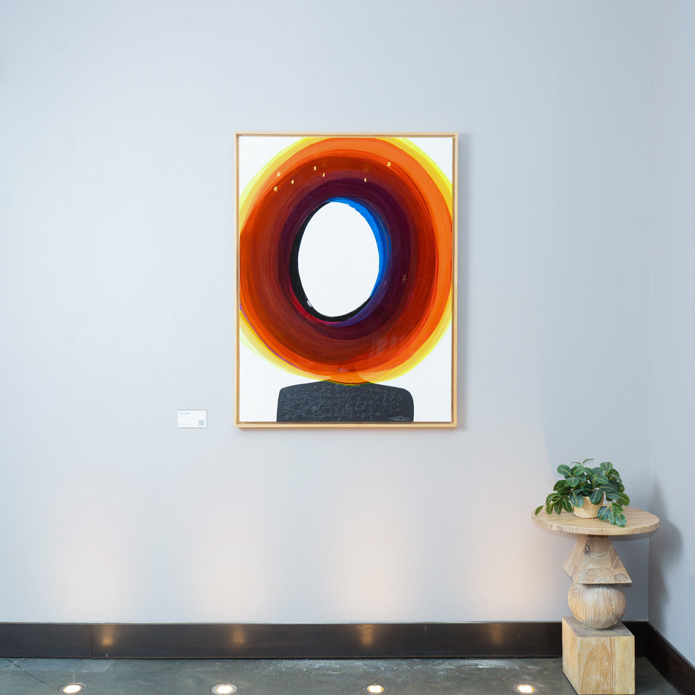 Abstract painting with concentric circles in red, orange, and blue hues on a gallery wall; a small round table with a plant is on the right side.