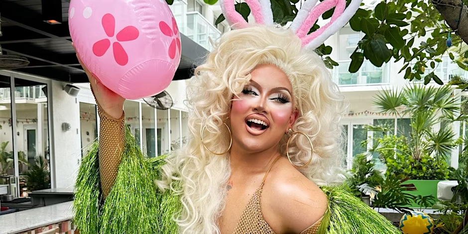 Person in a sparkly green outfit and bunny ears holds a pink, flower-decorated egg balloon, smiling outdoors under leafy canopy.