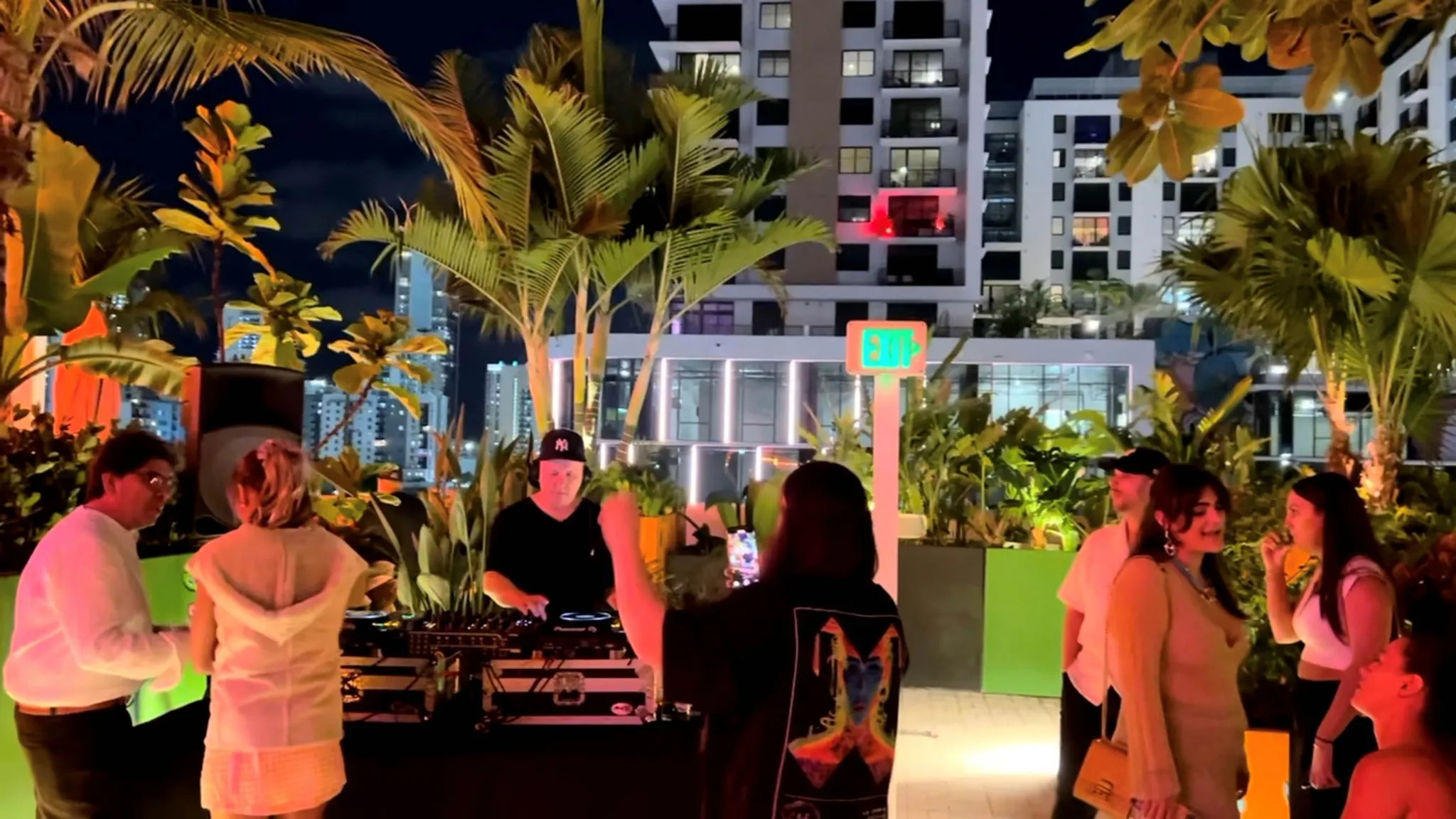 People enjoy music from a DJ at a rooftop party at night, surrounded by plants and city buildings.