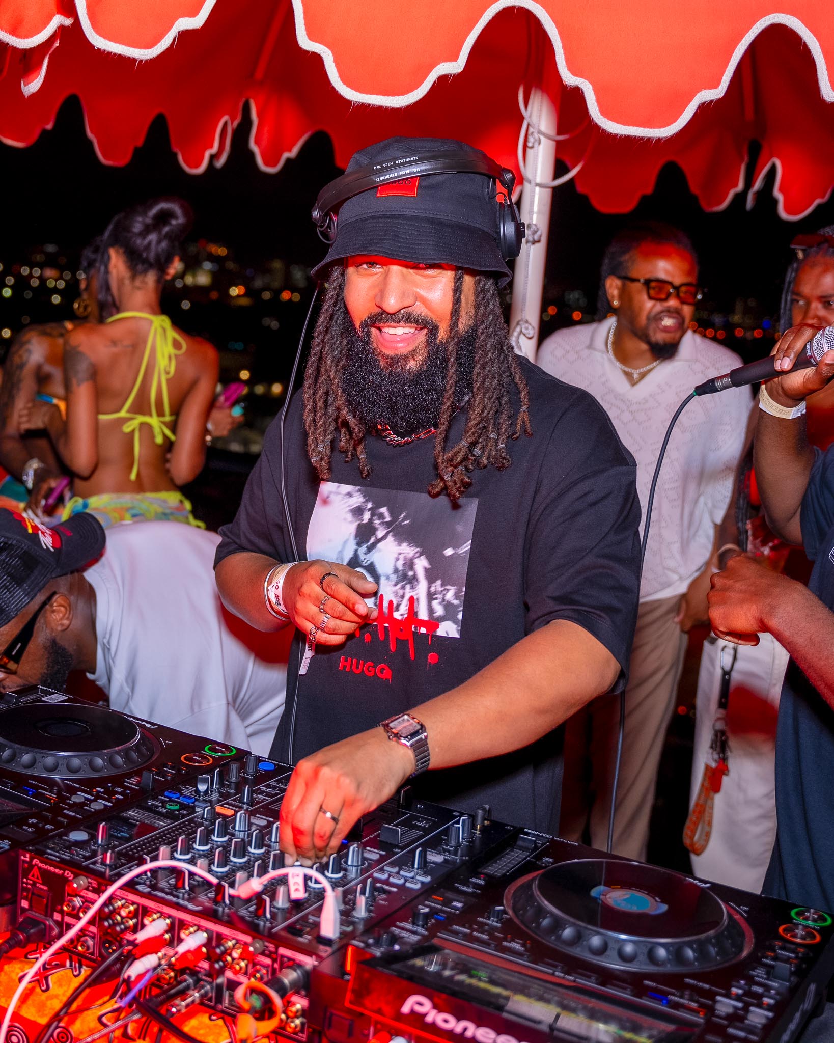 DJ with long hair and beard playing music on a Pioneer mixer under a red umbrella at a lively outdoor event. People are socializing and dancing in the background.
