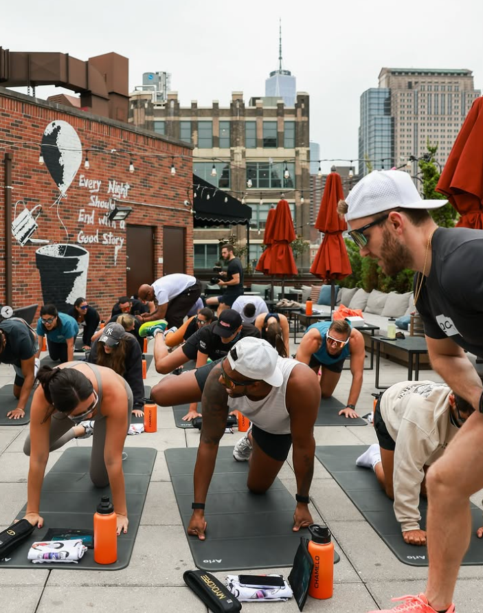 A group of people doing a guided yoga session on a rooftop with city buildings in the background.