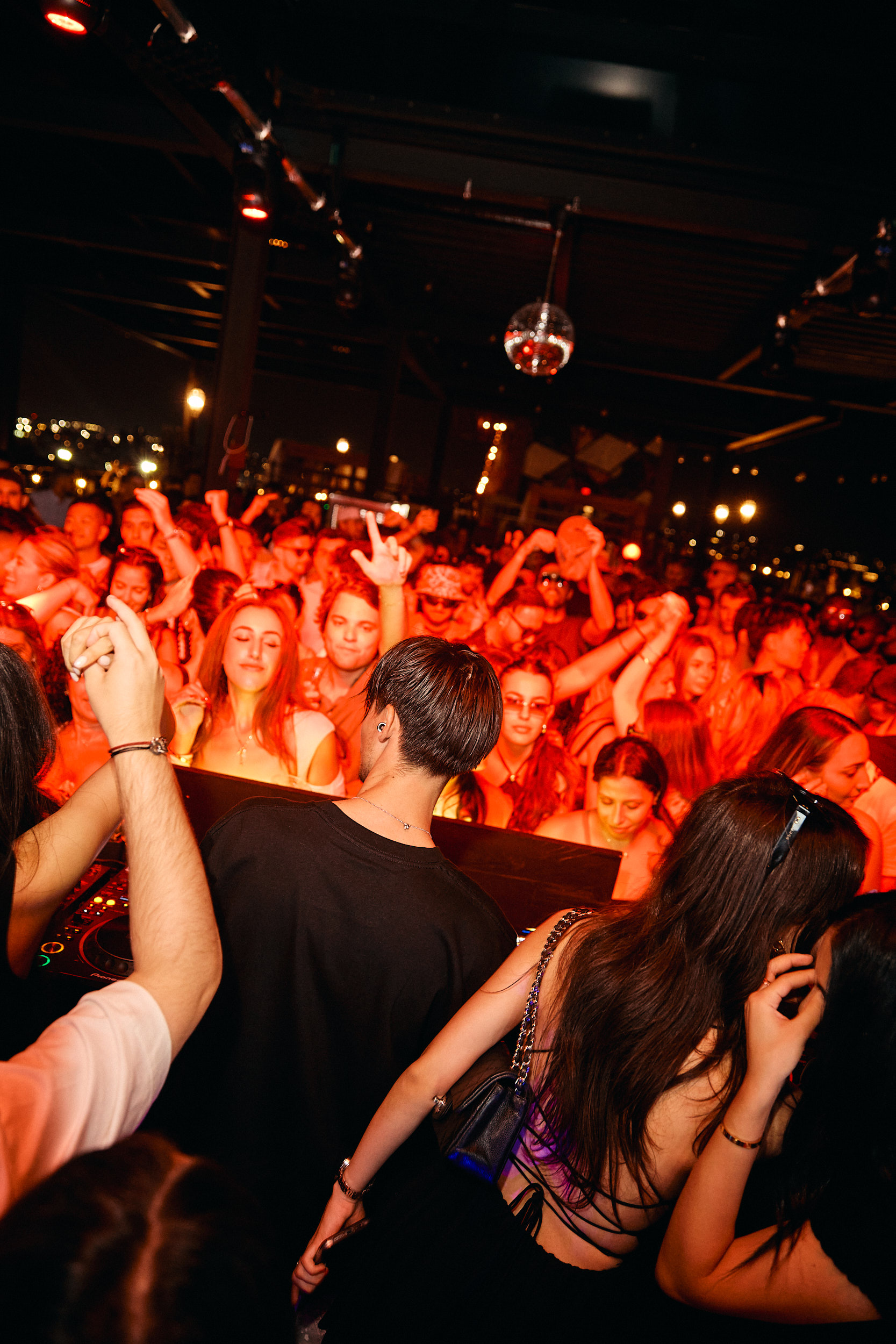 A crowd of people dances and raises their hands at a nightclub or concert with red lighting, seen from behind a DJ at the booth.