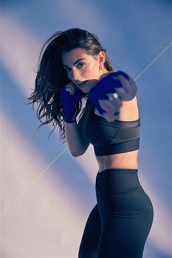 Woman in athletic wear, with blue hand wraps, poses in a boxing stance, focusing on her outstretched fist against a blue-toned background.