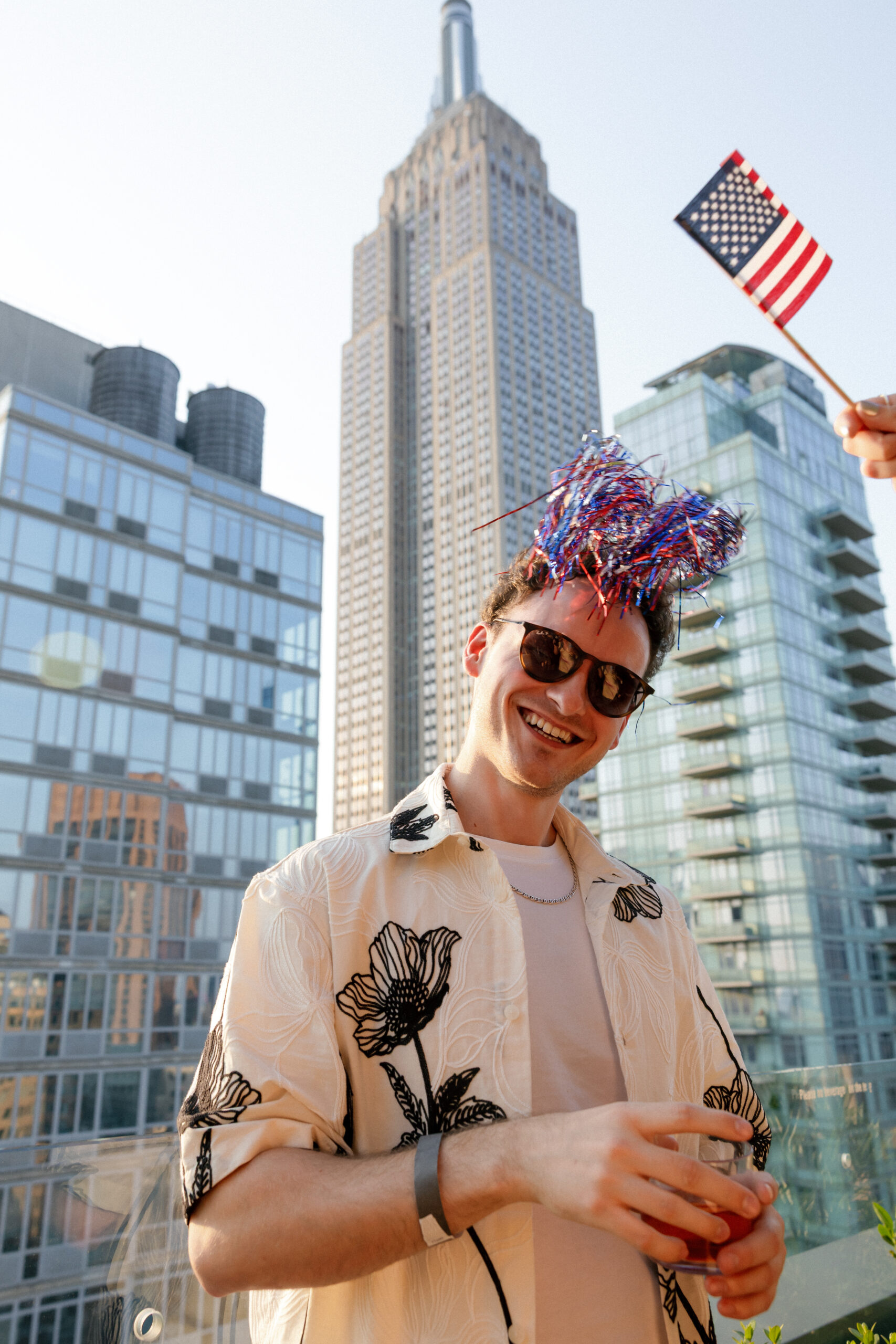 Man wearing sunglasses and a festive headband stands on a rooftop holding a drink, with the Empire State Building and modern glass buildings in the background; a small American flag is visible.