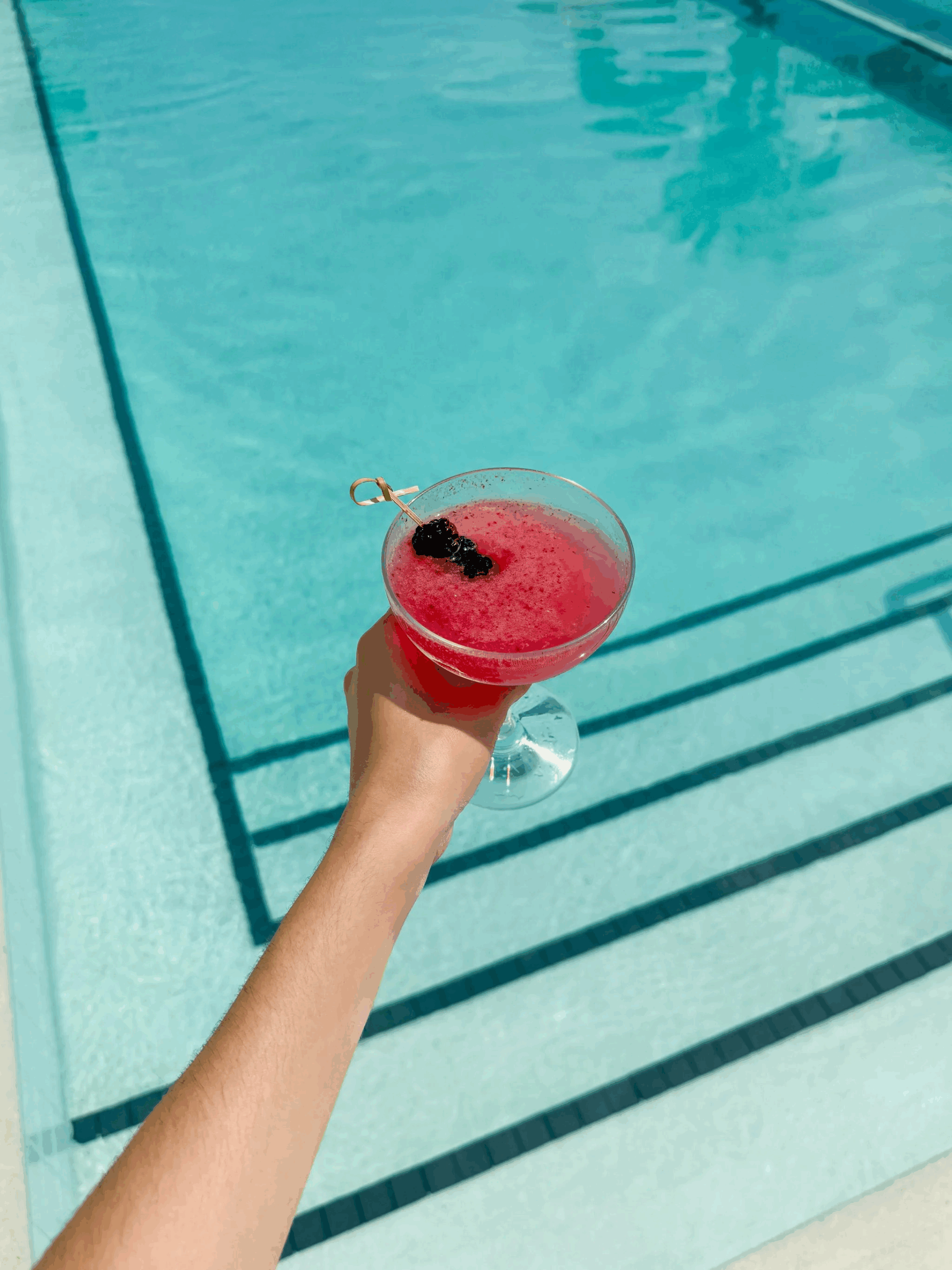 A hand holds a red cocktail with a blackberry garnish above swimming pool steps on a sunny day.