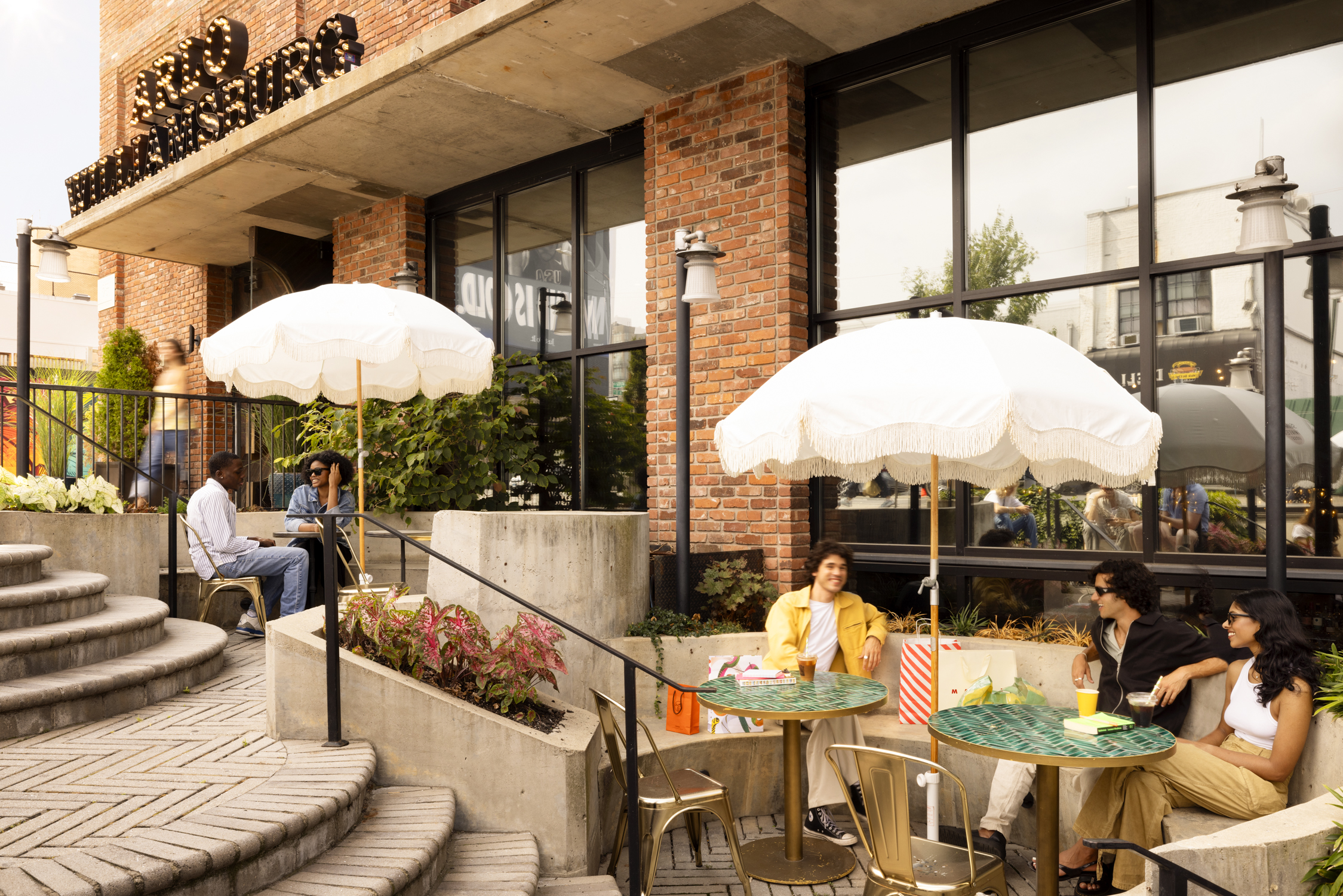 People sit at outdoor tables with drinks under white umbrellas outside a brick building on a sunny day.