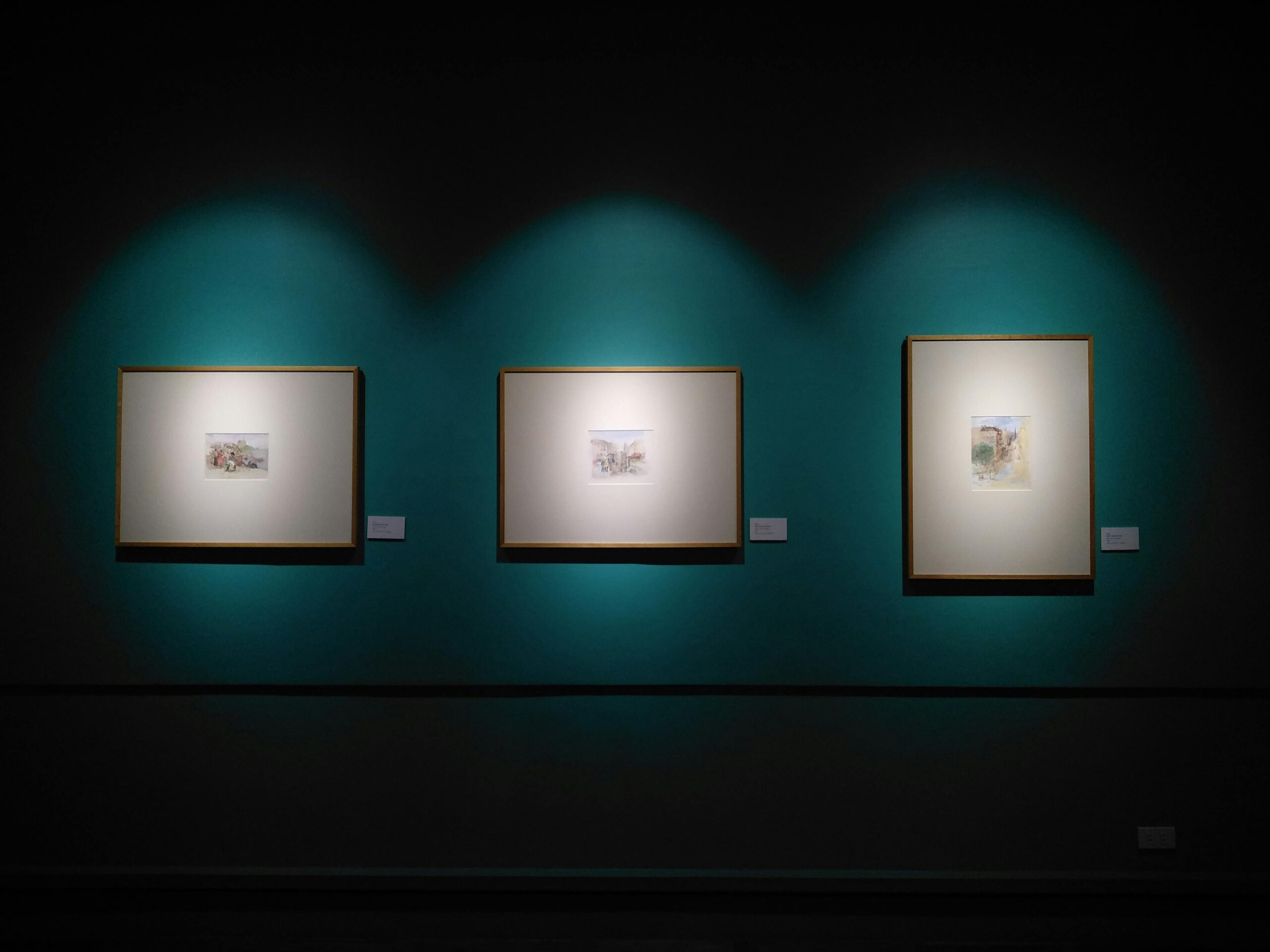 Three framed artworks, each illuminated by individual spotlights, are displayed on a teal wall in a dimly lit gallery reminiscent of the stylish museums in Soho NYC.