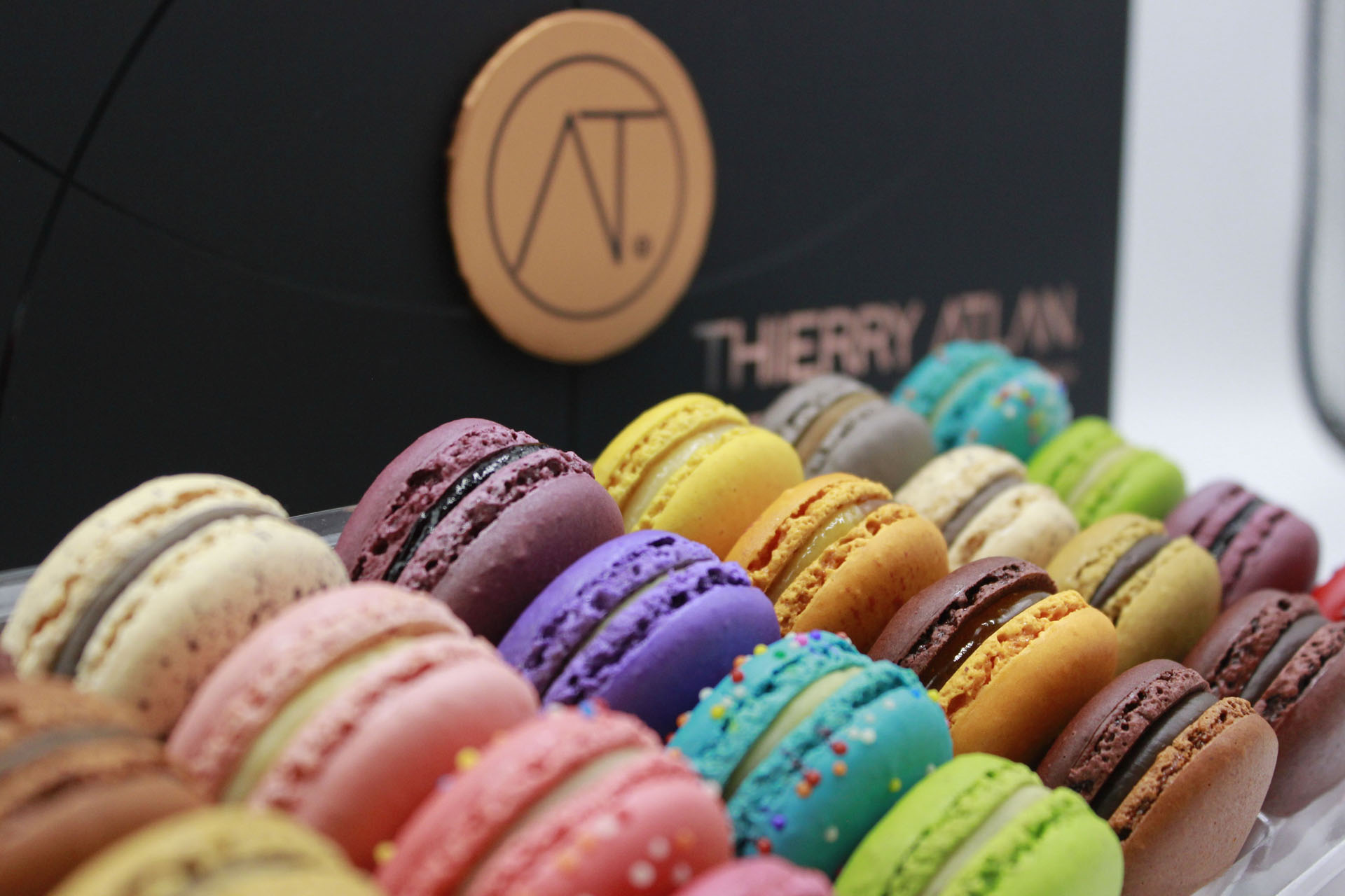A close-up of assorted colorful macarons arranged in rows, with a Thierry Atlan branded box in the background.