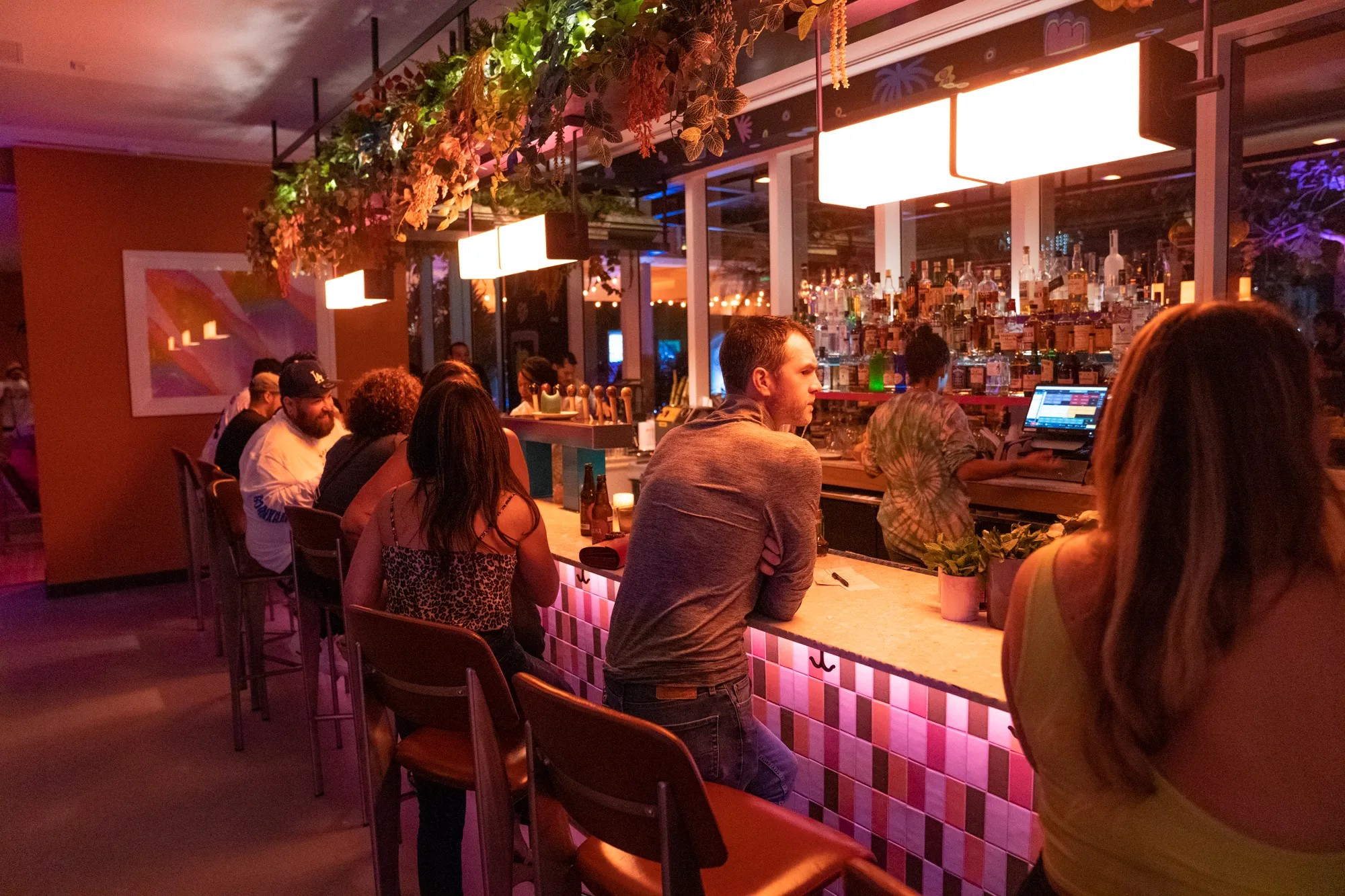 People sit and converse at a bar counter in a warmly lit, modern bar with plants and bottles displayed behind the counter.