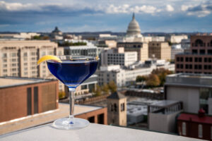 A blue cocktail with a lemon twist sits on a rooftop ledge of a rooftop bar in Washington DC, overlooking city buildings and the U.S. Capitol dome in the background.