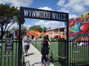 Entrance gate to Wynwood Walls with people taking photos, vibrant Wynwood Walls street art murals in the background, and a clear blue sky above.