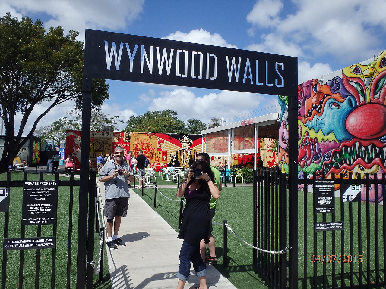 Entrance gate to Wynwood Walls with people taking photos, vibrant Wynwood Walls street art murals in the background, and a clear blue sky above.