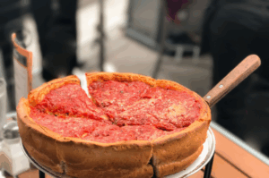 A deep-dish pizza with a thick crust, topped with tomato sauce and cheese, sits on a metal tray with a wooden-handled server on a table.