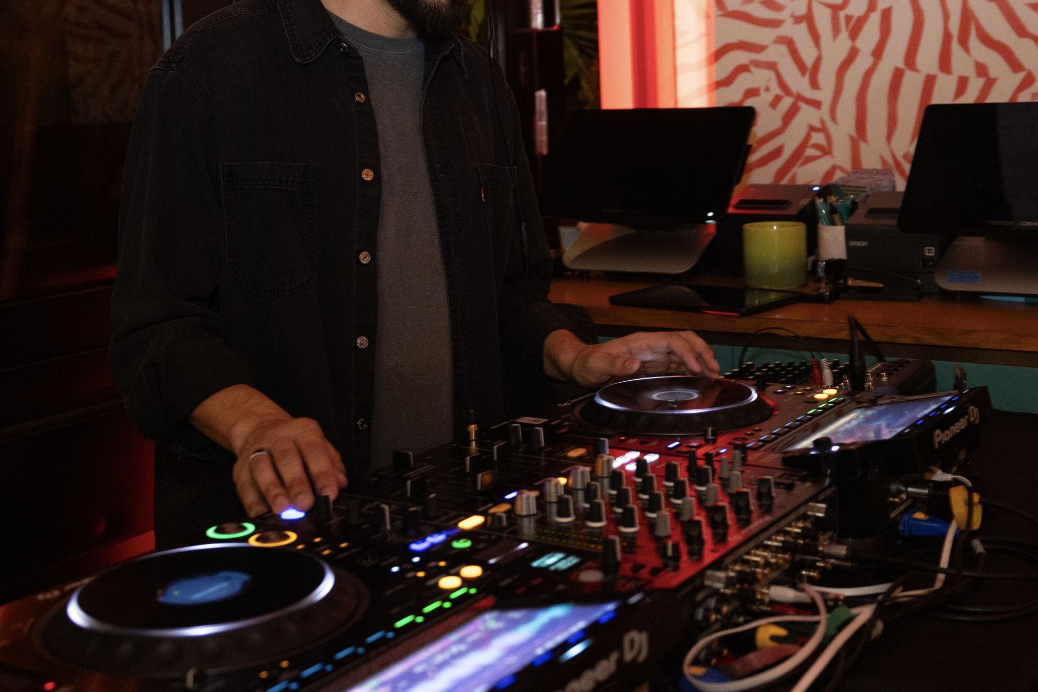 A person using DJ equipment, adjusting controls on a mixer and turntables in a dimly lit room with computers and office supplies in the background.
