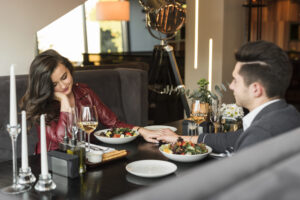 A woman and man sit at a restaurant table holding hands, with salads, wine, and candles in front of them. The woman is smiling and looking down during an intimate evening of fine dining in Manhattan New York.