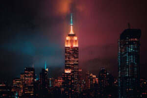 The Empire State Building in NYC is illuminated at night, surrounded by city skyscrapers under a cloudy, colorfully lit sky.