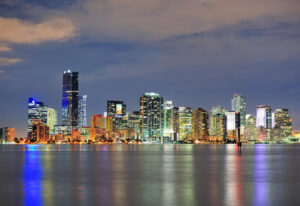 Experience the vibrant city skyline at night—a highlight of what to see in downtown Miami—with tall buildings illuminated, their lights reflecting on the calm water below a partly cloudy sky.
