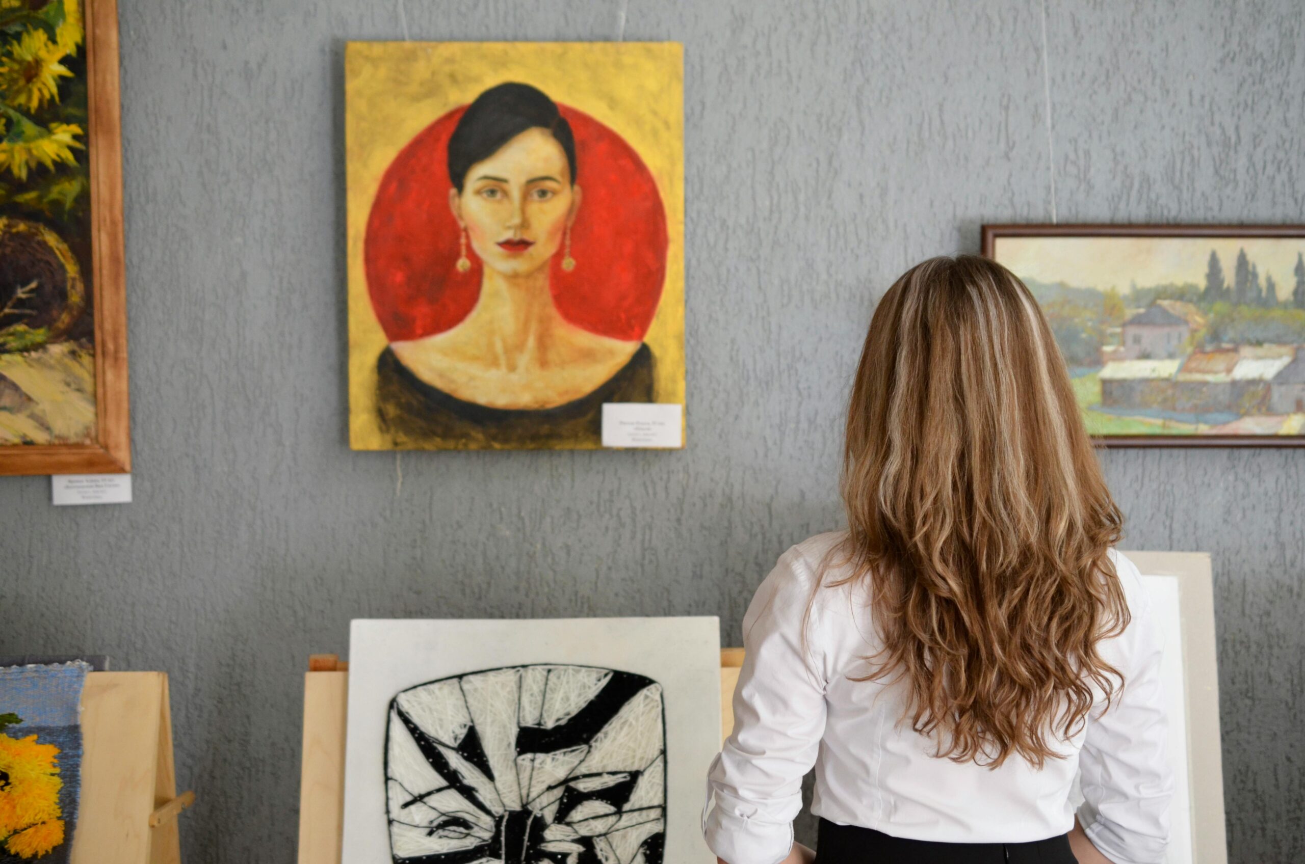 A woman with long, wavy hair stands in front of paintings displayed on a gallery wall at one of the art galleries in Williamsburg Brooklyn, admiring a portrait of a woman with a red circle background.