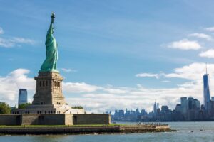 The Statue of Liberty stands on Liberty Island, one of the iconic NYC landmarks, with the New York City skyline and One World Trade Center visible in the background under a blue sky.