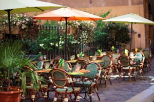 Outdoor café seating with multiple tables and chairs, green plants, and umbrellas set on a cobblestone patio beside a building and iron fence—perfect for relaxed Wynwood dining.