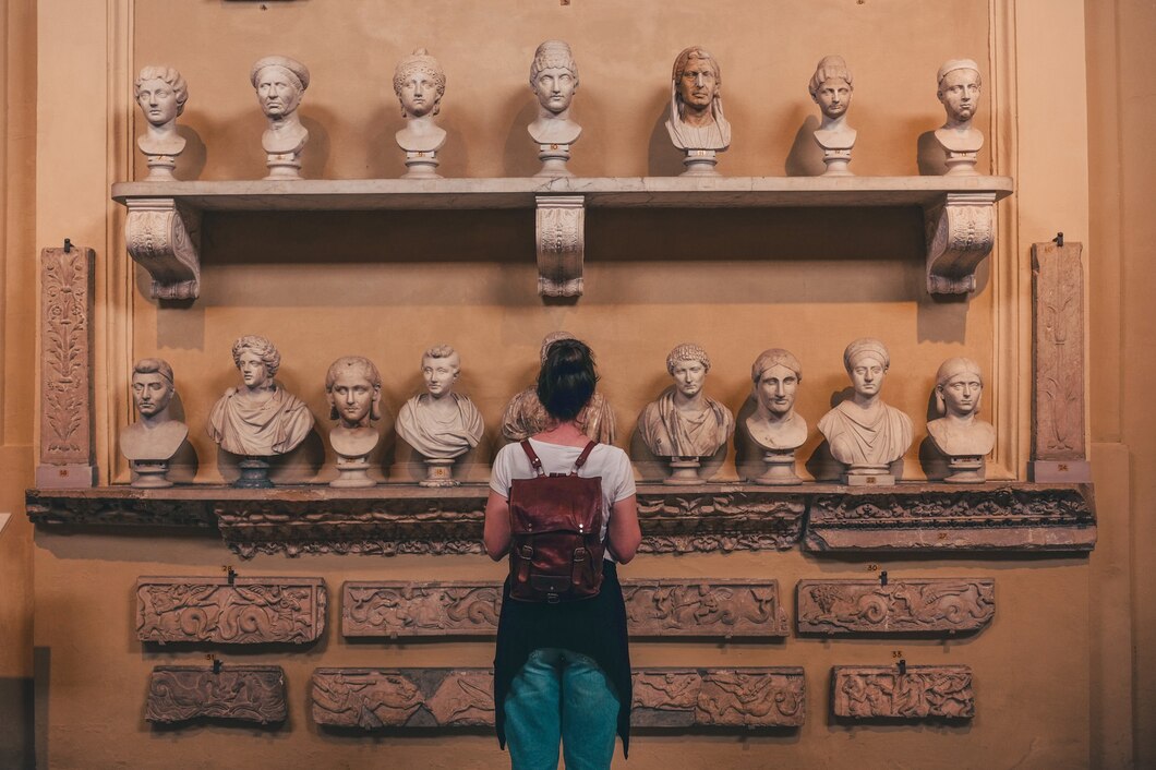 A person with a backpack stands in front of two shelves displaying ancient marble busts in an NYC museum or gallery with a yellow wall.