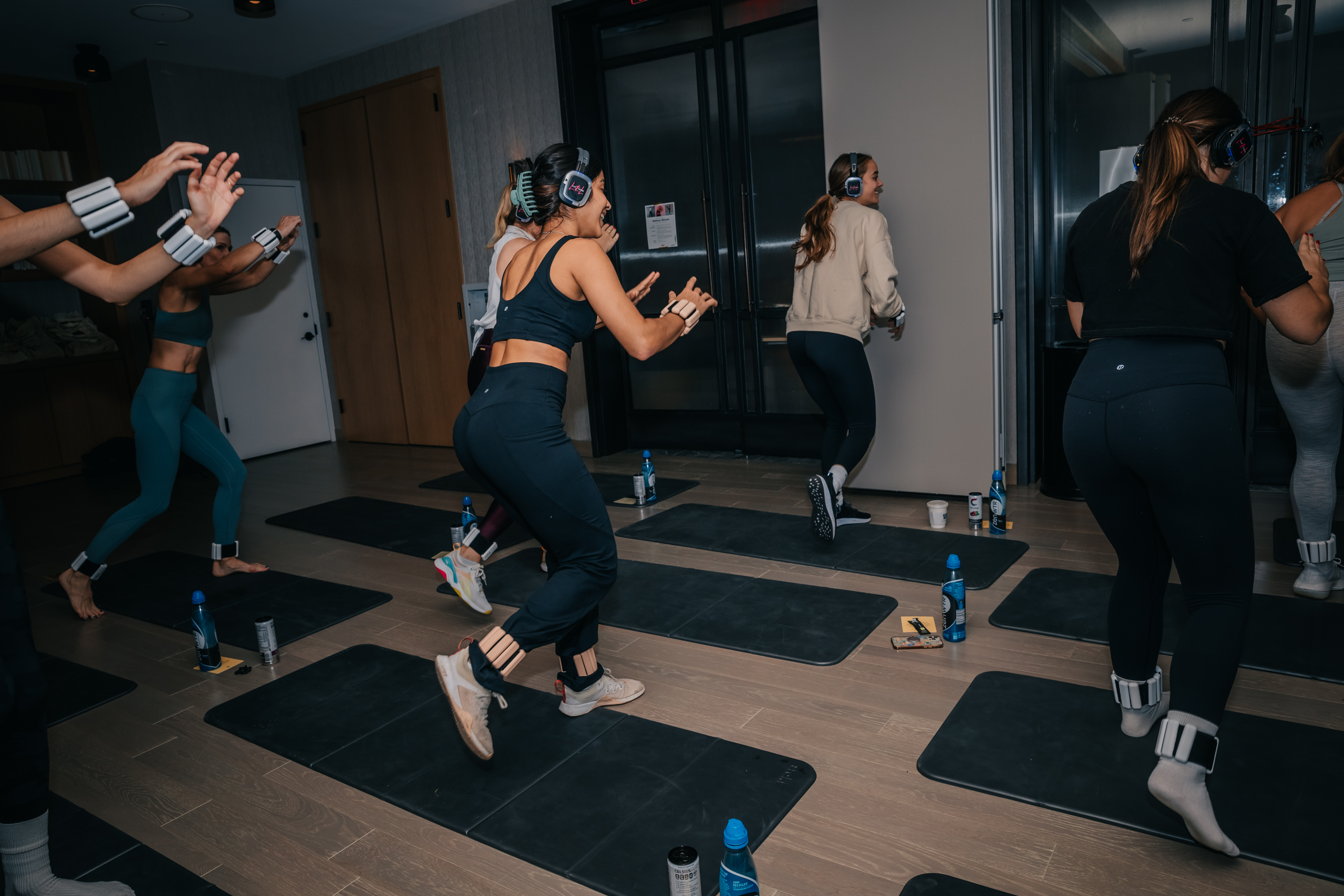A group of people wearing headphones and ankle weights exercise on mats in a fitness studio, with water bottles placed beside them.