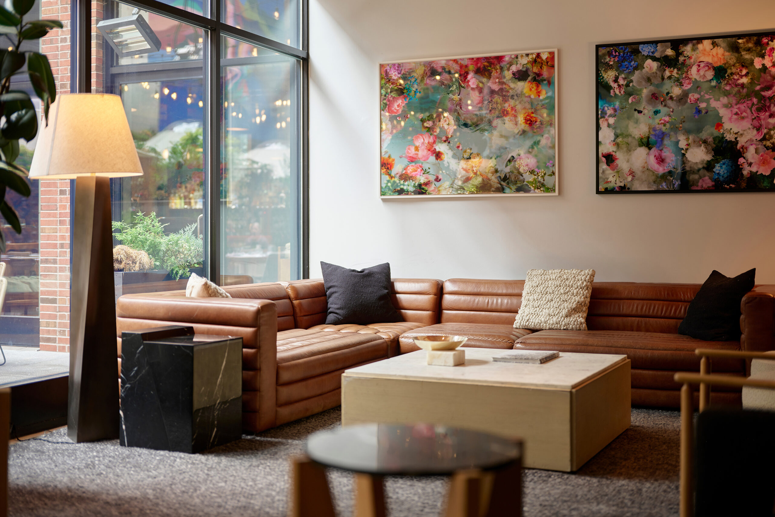 Modern lounge area with brown leather sectional sofa, black and beige cushions, light wood coffee table, abstract floral wall art, and floor-to-ceiling windows.