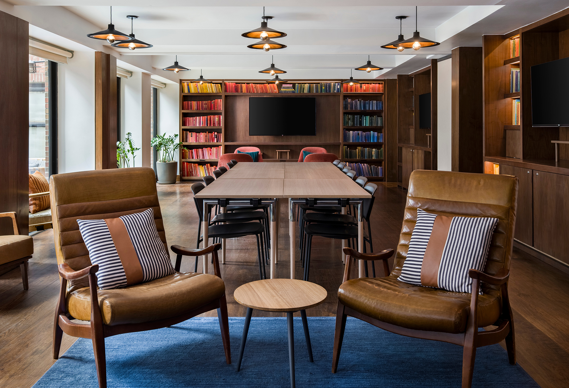 Modern meeting room with two leather armchairs, a small round table, long conference tables, black chairs, bookshelves, and mounted TVs. Warm lighting and wood accents throughout.