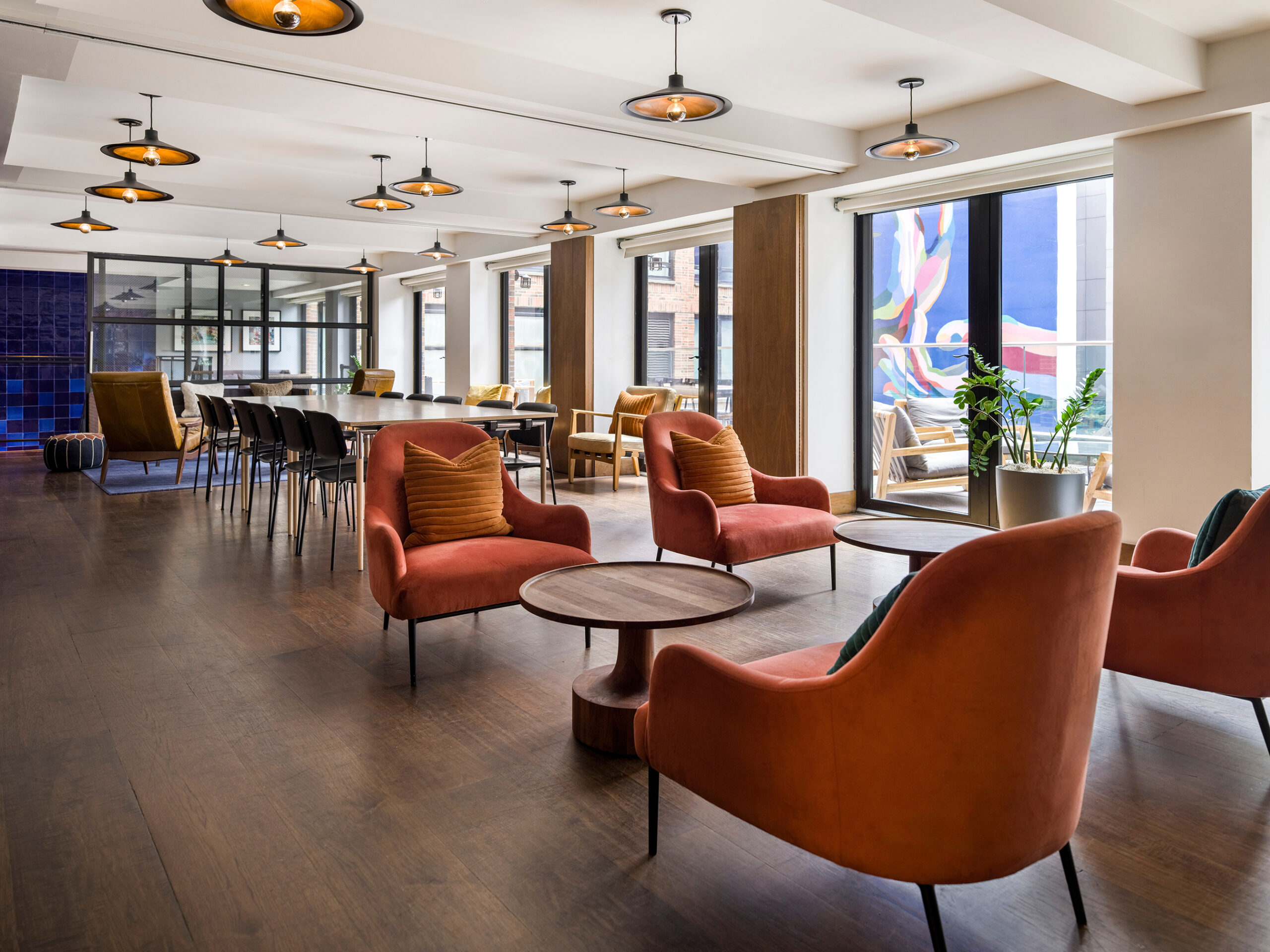 Modern lounge area with orange armchairs, round tables, long communal tables, and large windows letting in natural light. Contemporary lighting fixtures hang from the ceiling.