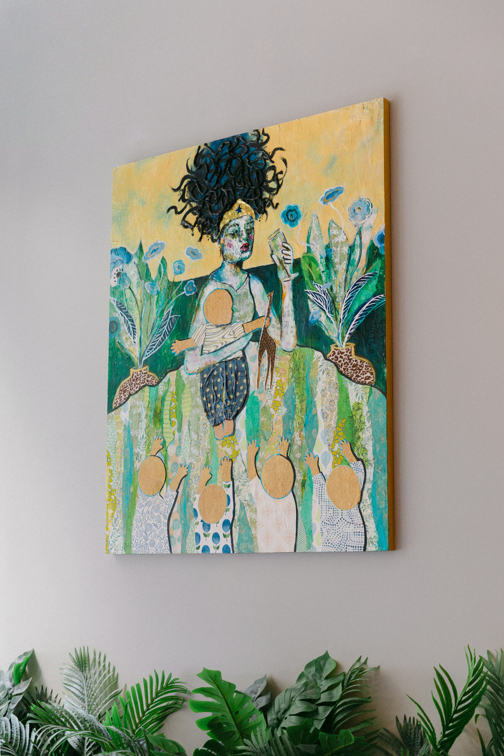 A colorful painting of a crowned woman with dark curly hair, greenery, and abstract figures, hangs on a light wall above potted green plants.