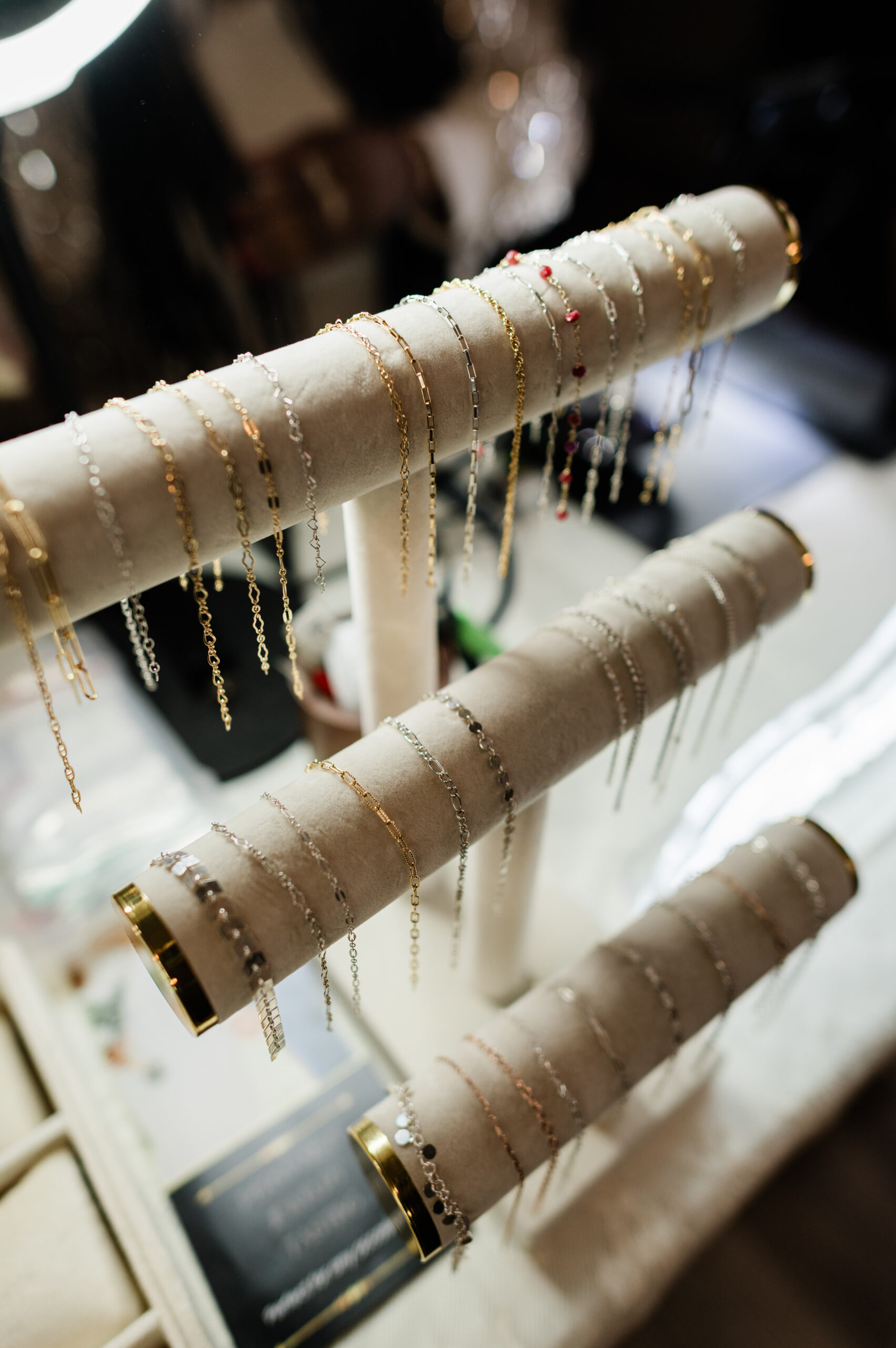Three cushioned display rods hold an assortment of delicate gold, silver, and beaded chain bracelets, arranged neatly in rows on a jewelry stand.