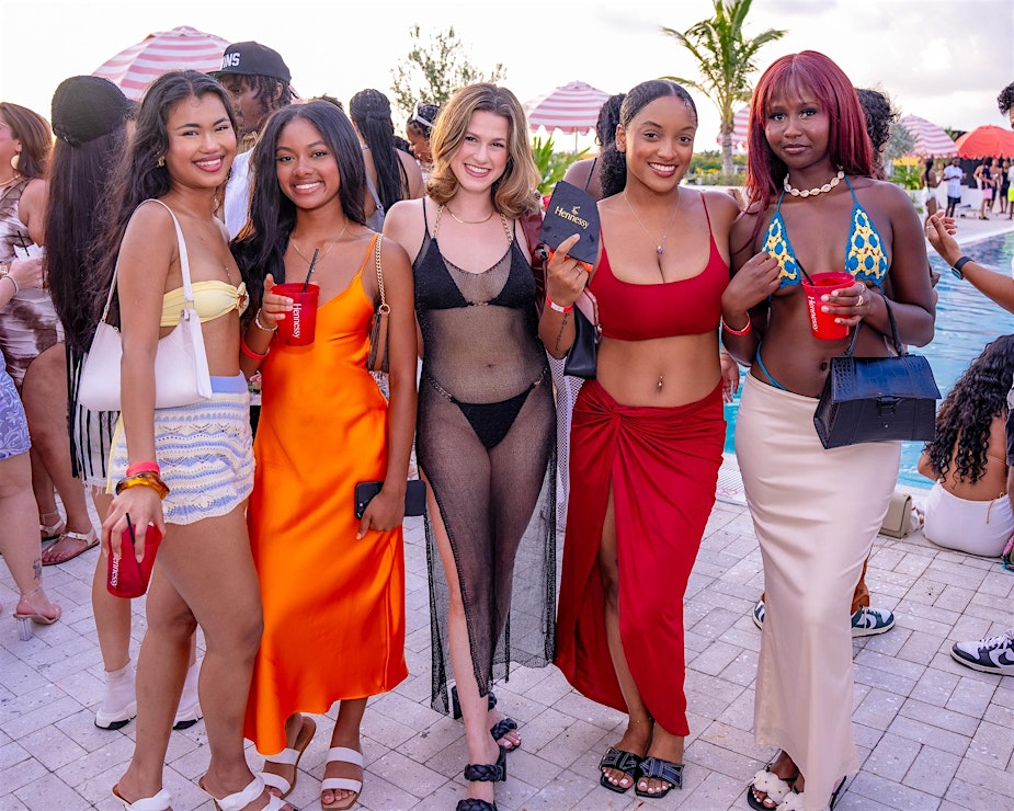 Five women pose together by a pool, dressed in colorful summer outfits and holding red cups, with other people and umbrellas in the background.