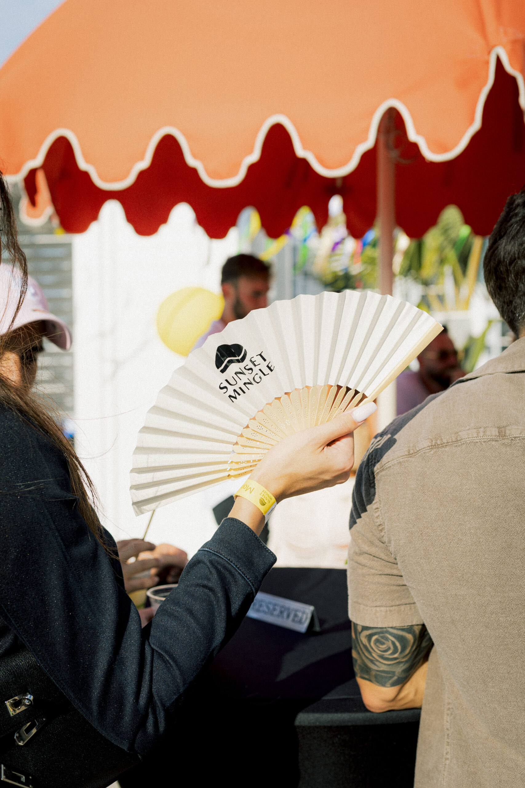 Person holding a white fan with "Sunset Mingle" text, standing under a red umbrella at an outdoor event.