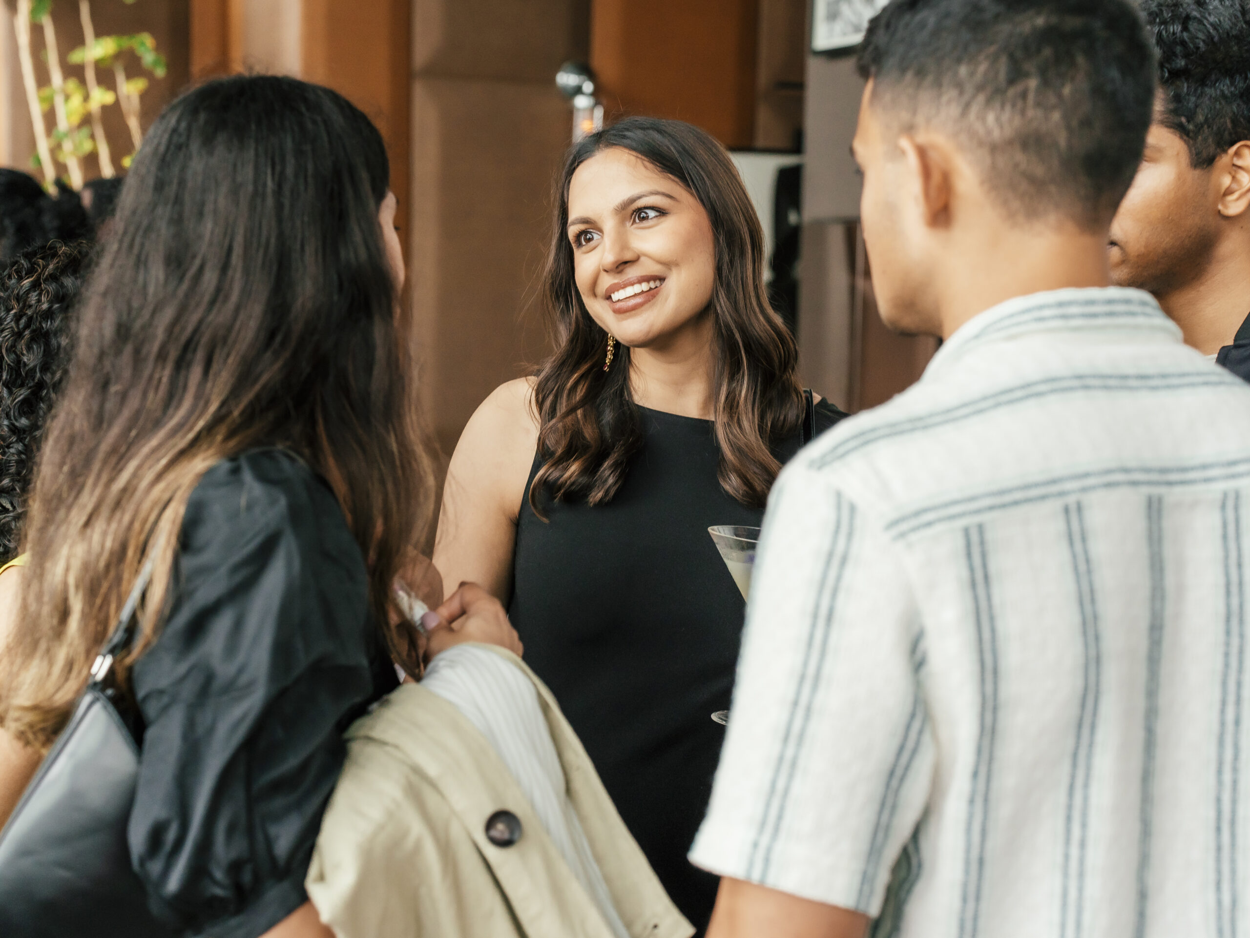 A woman holding a drink smiles while talking with three people at an indoor social gathering.