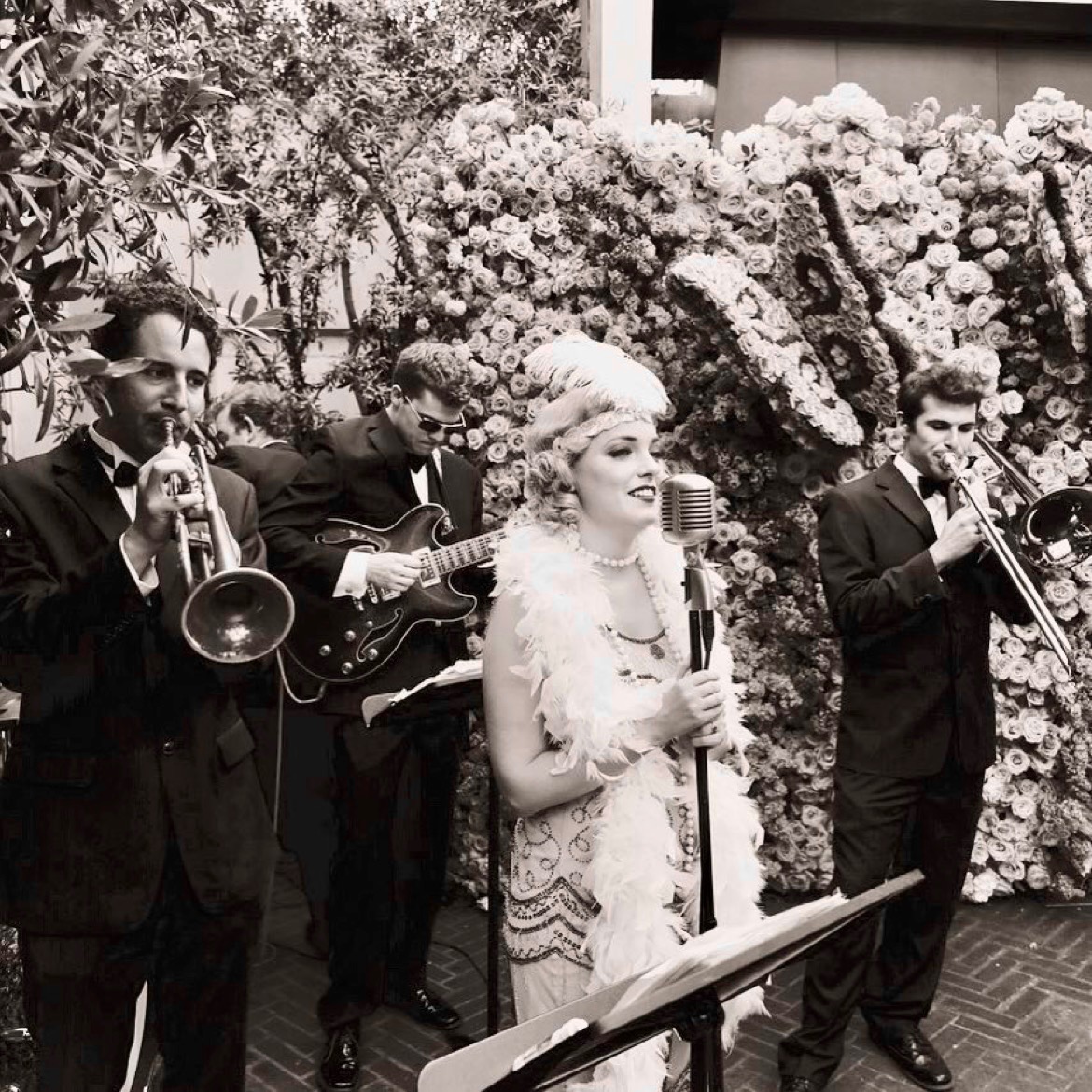 A jazz band dressed in vintage attire performs outdoors; a woman sings at a microphone while three men play trumpet, guitar, and trombone in front of a wall of flowers.