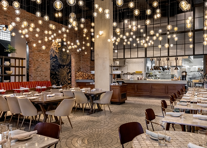 A modern restaurant dining area with round tables, chairs, exposed brick walls, open kitchen, and numerous hanging globe lights from the ceiling.