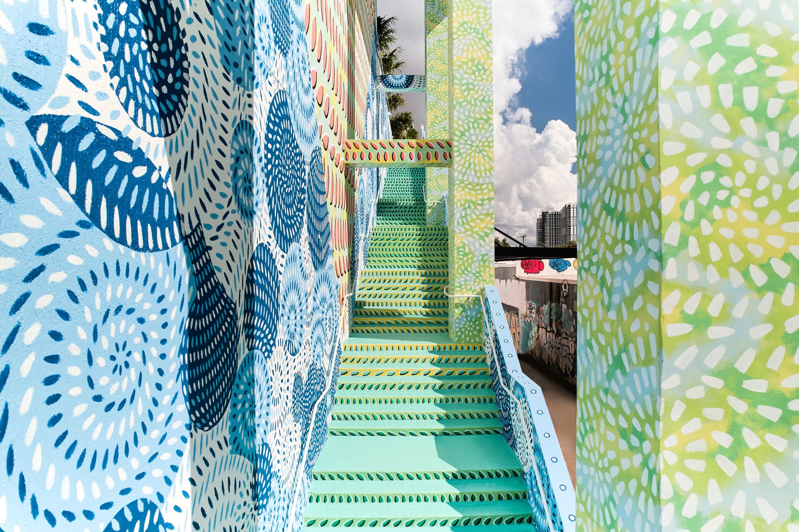 Colorful outdoor staircase decorated with swirling blue, green, and white patterns, framed by patterned walls, with city buildings and sky visible in the background.