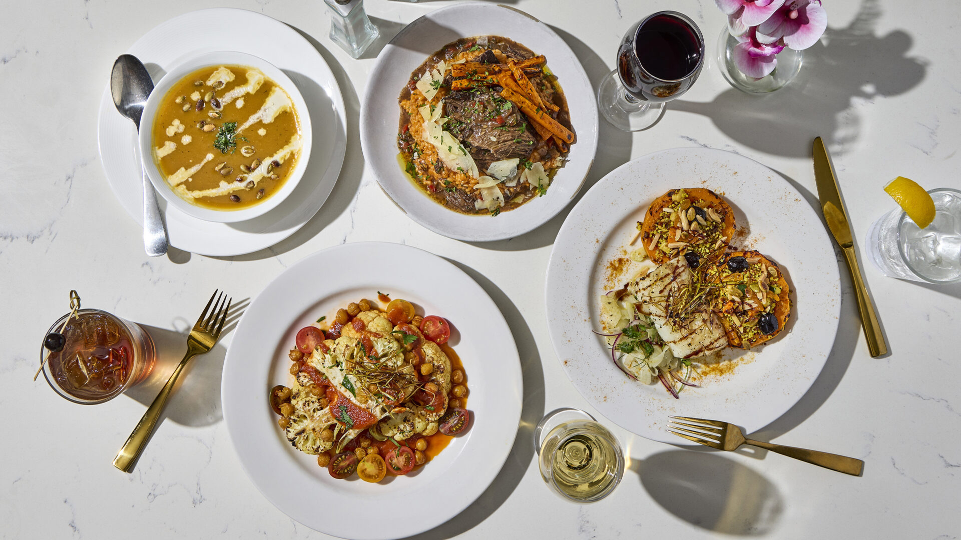 Four plated gourmet dishes with drinks, including soup, steak with vegetables, pasta, and a vegetable dish, arranged on a white marble table with gold cutlery and flowers.