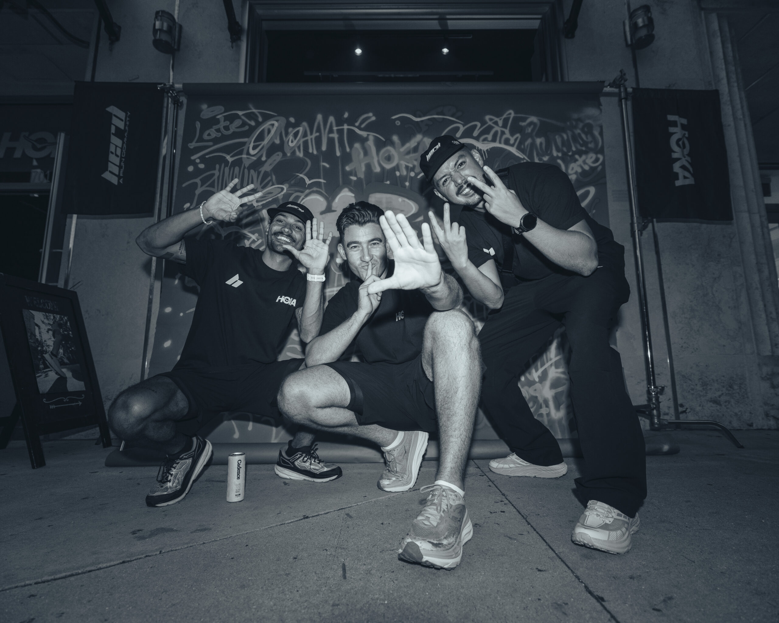 Three men in athletic wear crouch and pose playfully in front of a graffiti-covered backdrop on a sidewalk at night.