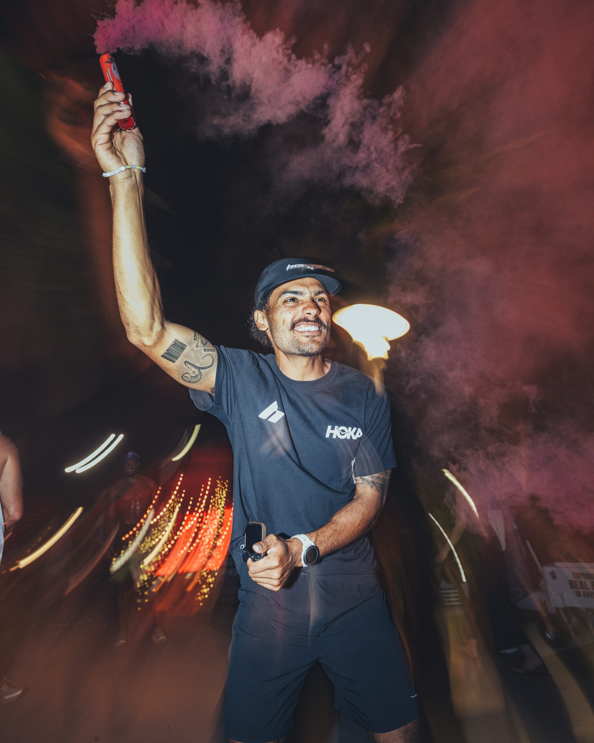A man in athletic clothing holds a red smoke flare aloft at night, surrounded by swirling pink smoke and blurred lights.