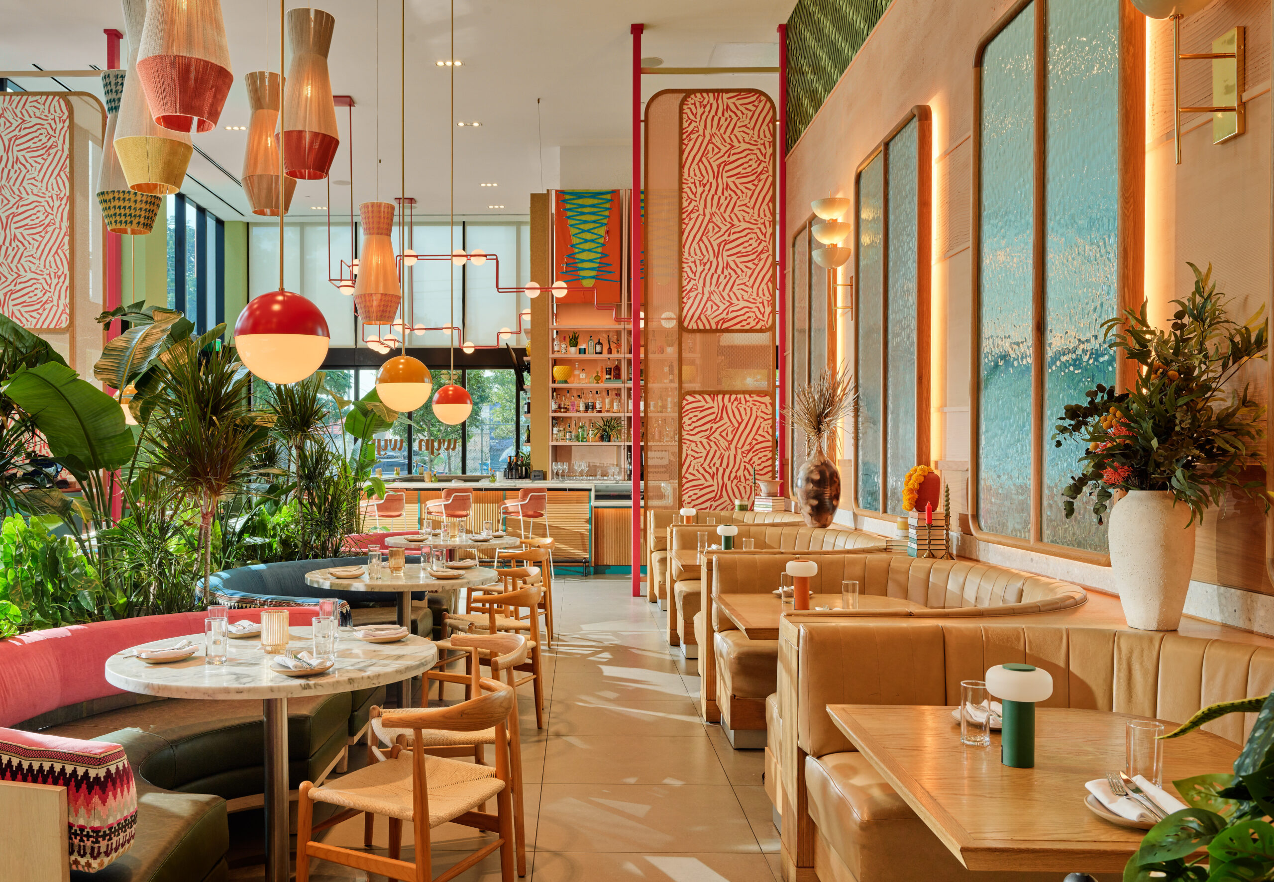 Modern restaurant interior with tan leather booths, round tables, wooden chairs, large plants, colorful light fixtures, and patterned wall art in a bright, spacious setting.