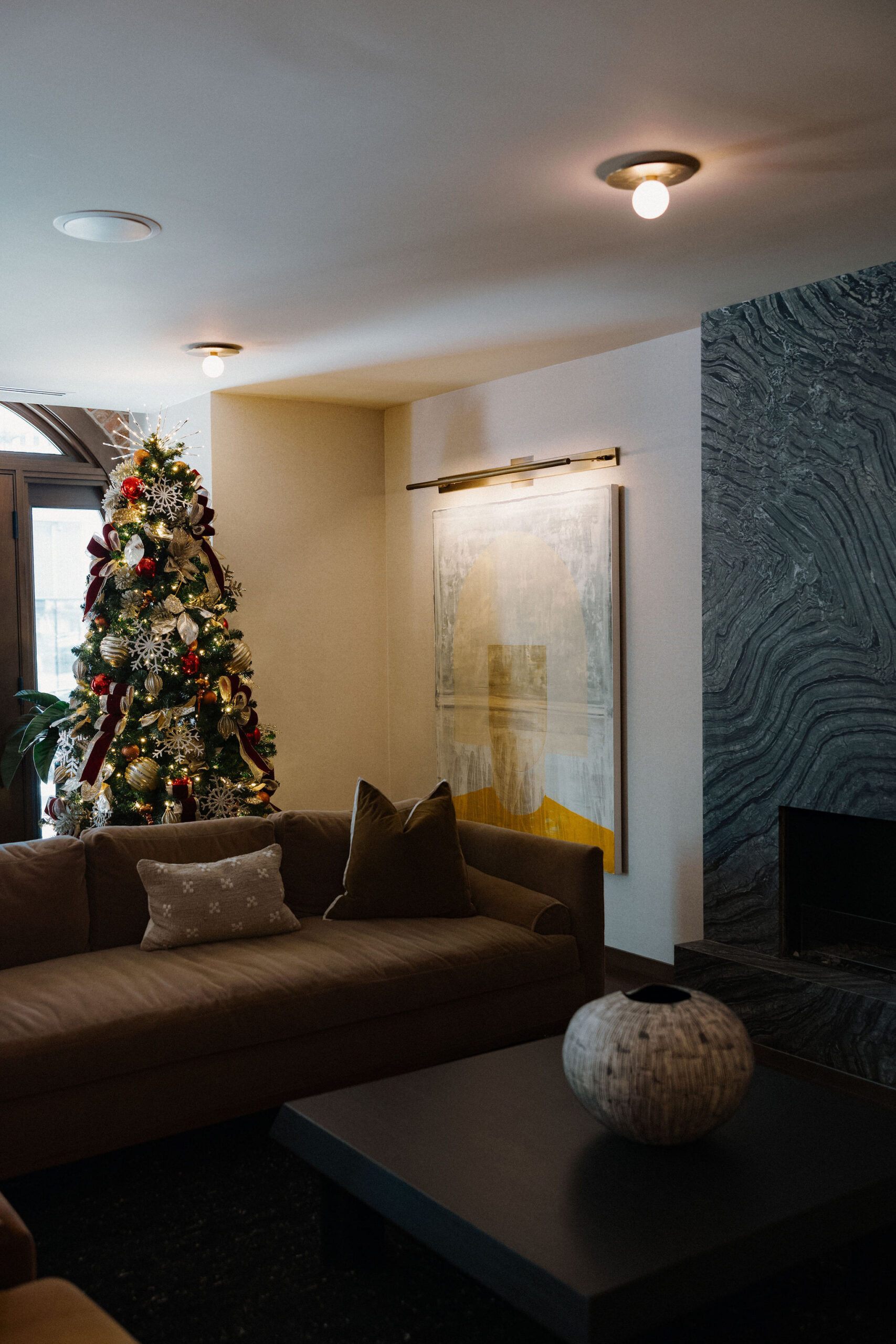 A living room with a decorated Christmas tree, a brown sofa with cushions, a large abstract painting, a dark coffee table, and a modern fireplace.
