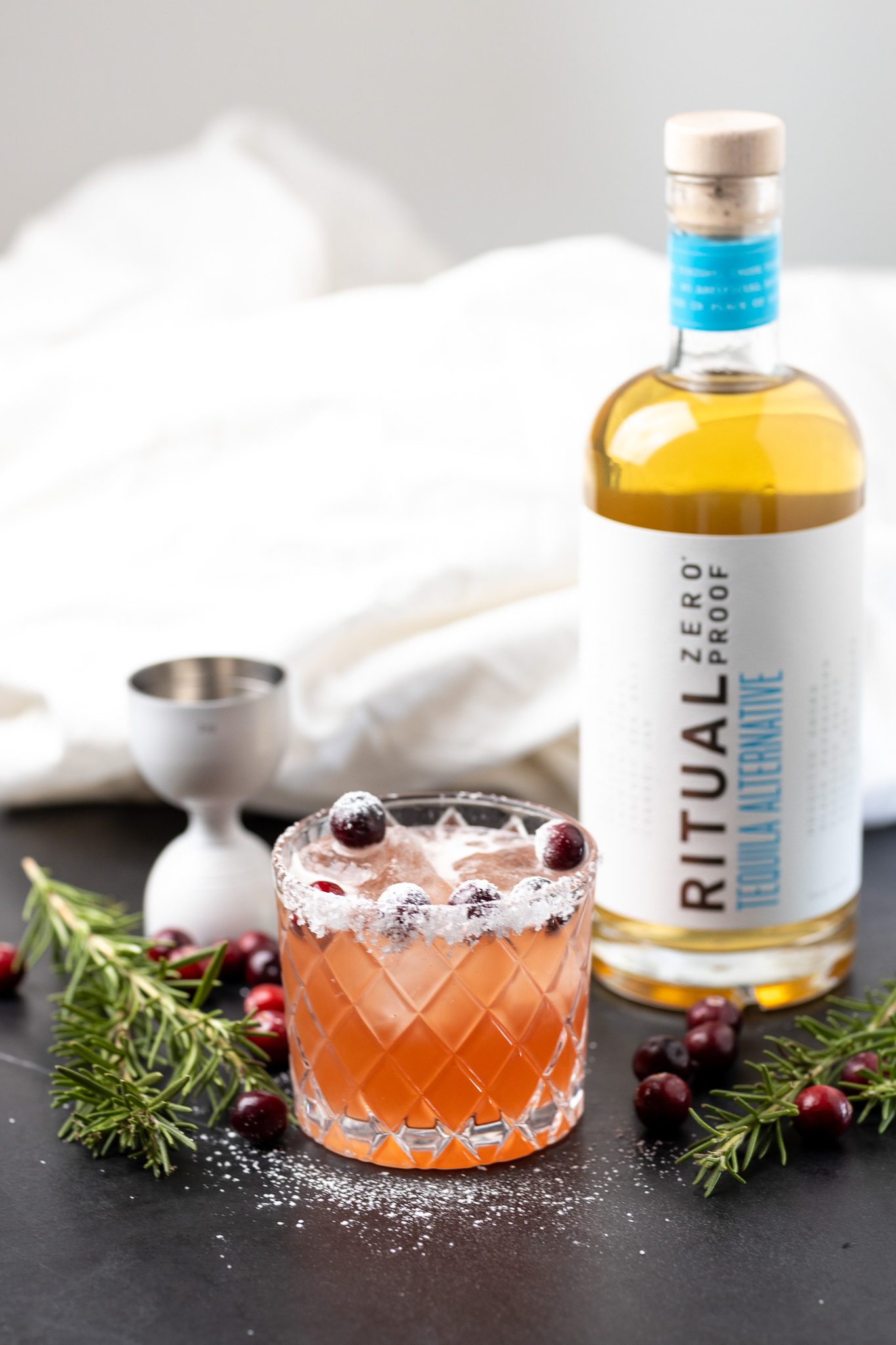 A glass of pink cocktail garnished with cranberries and rosemary sits next to a bottle labeled "Ritual Zero Proof Tequila Alternative" on a black surface.