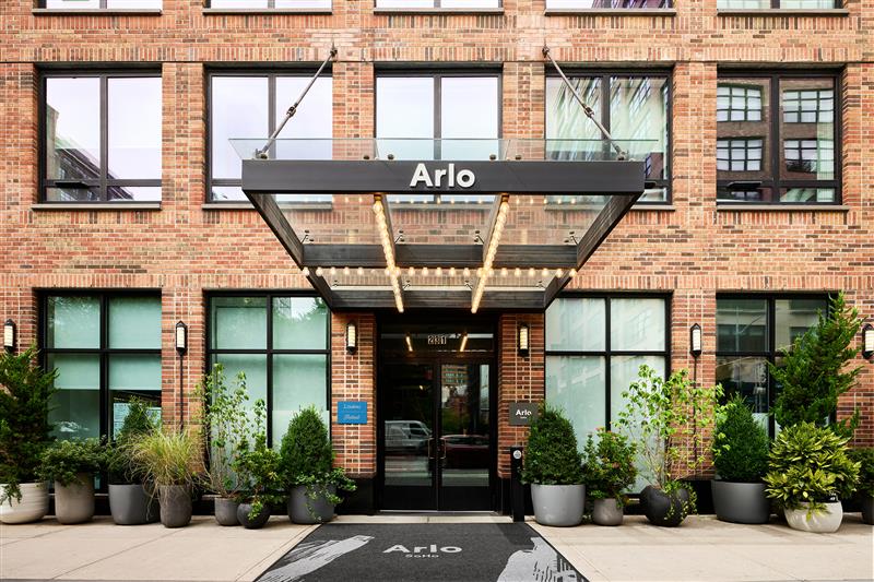 Exterior view of the Arlo hotel entrance with a glass canopy, potted plants, and large windows on a brick building facade.