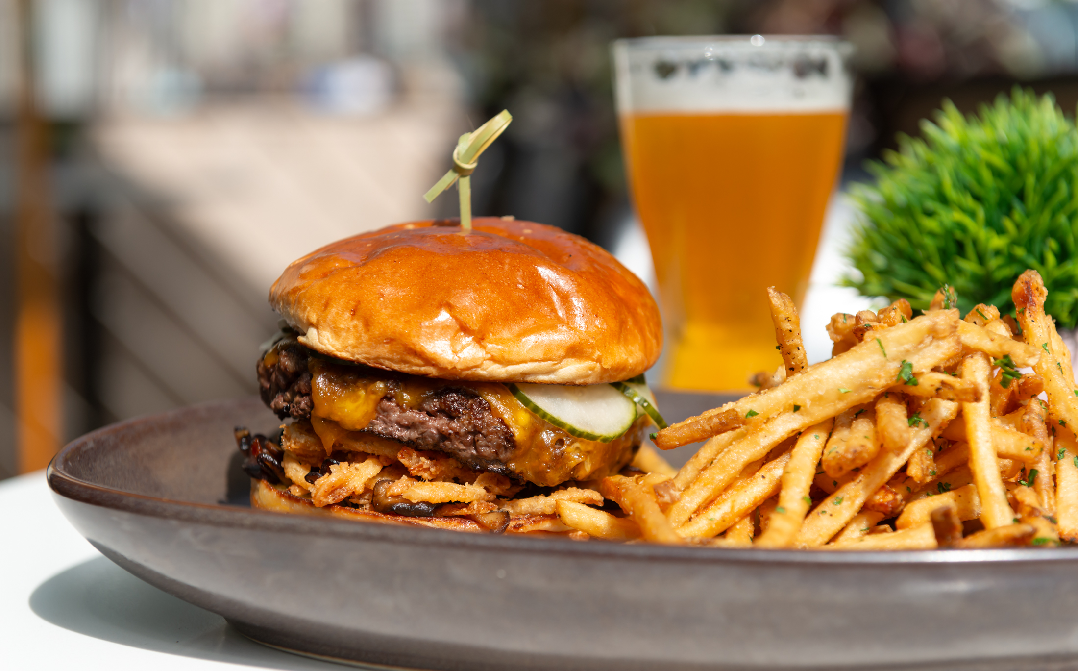 A cheeseburger with pickles and onions on a bun, served with seasoned French fries on a plate, and a glass of beer in the background.