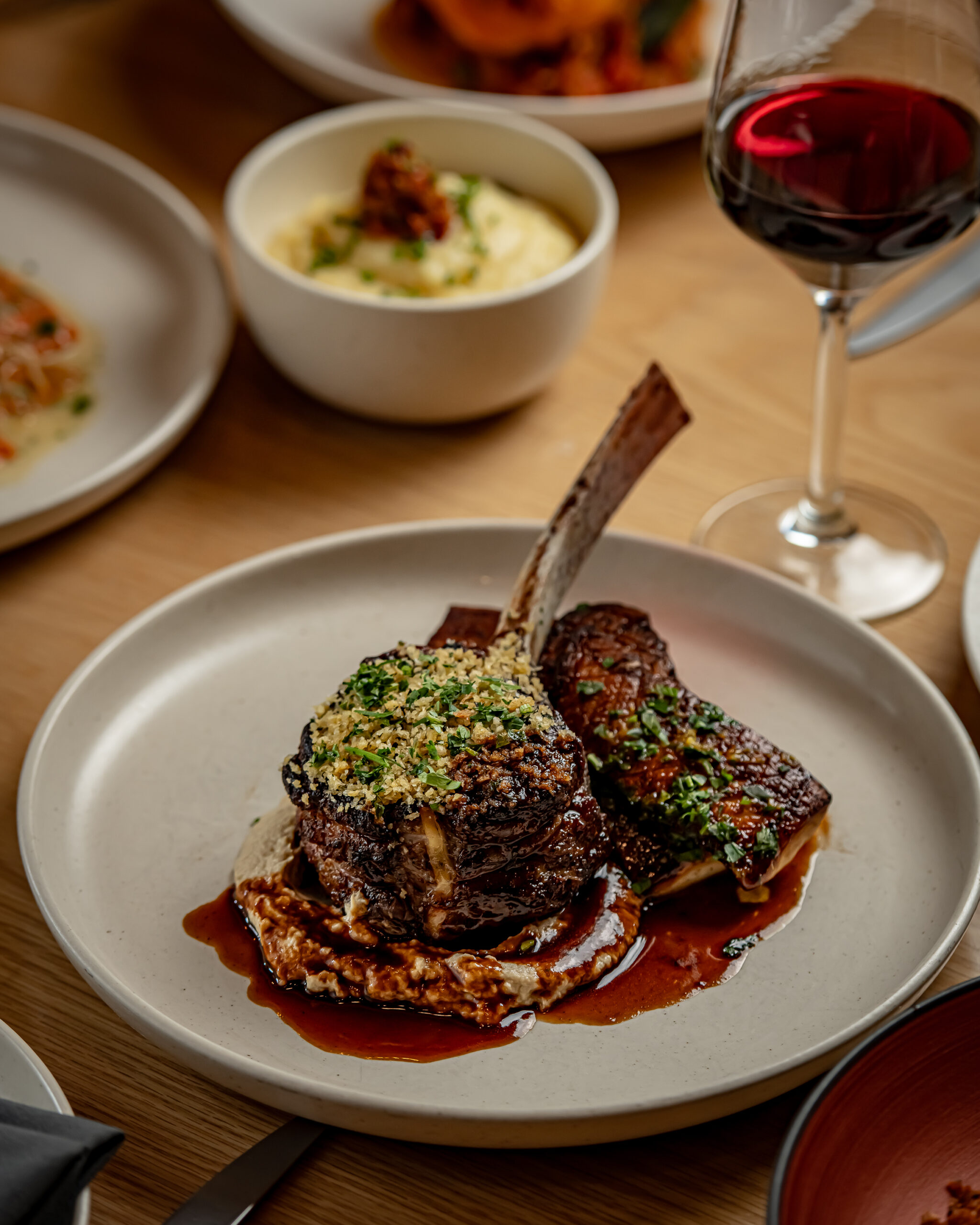 A plate with a tomahawk steak topped with herbs, served with sauce, mashed potatoes in a bowl, and a glass of red wine on a wooden table.