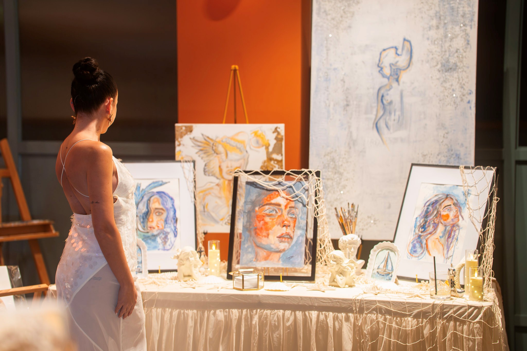 A woman in a white dress looks at a display table with framed paintings, art supplies, and sculptures against an orange and white background.