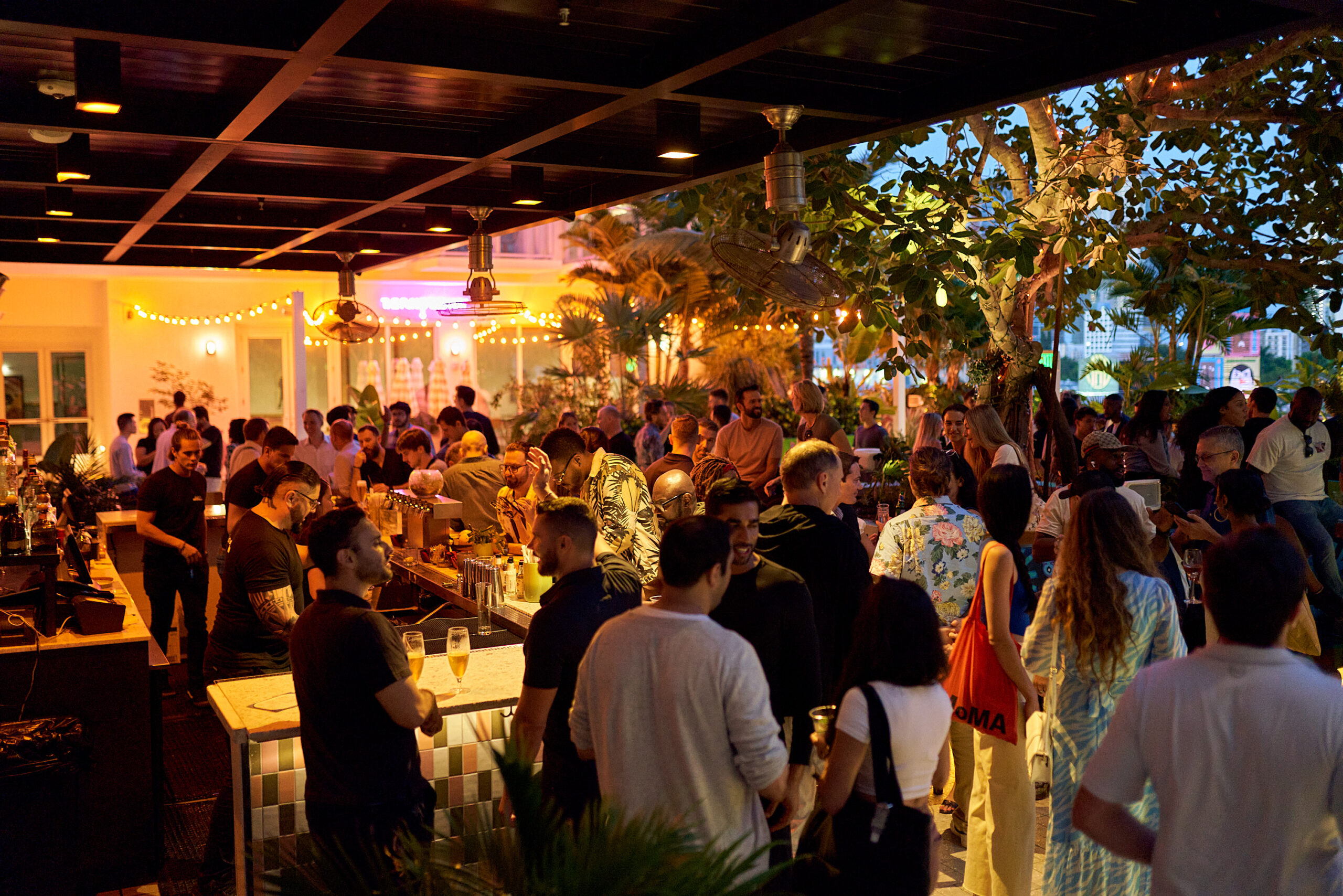 A lively crowd gathers at an outdoor bar in the evening, with people socializing, bartenders serving drinks, and string lights creating a festive atmosphere.