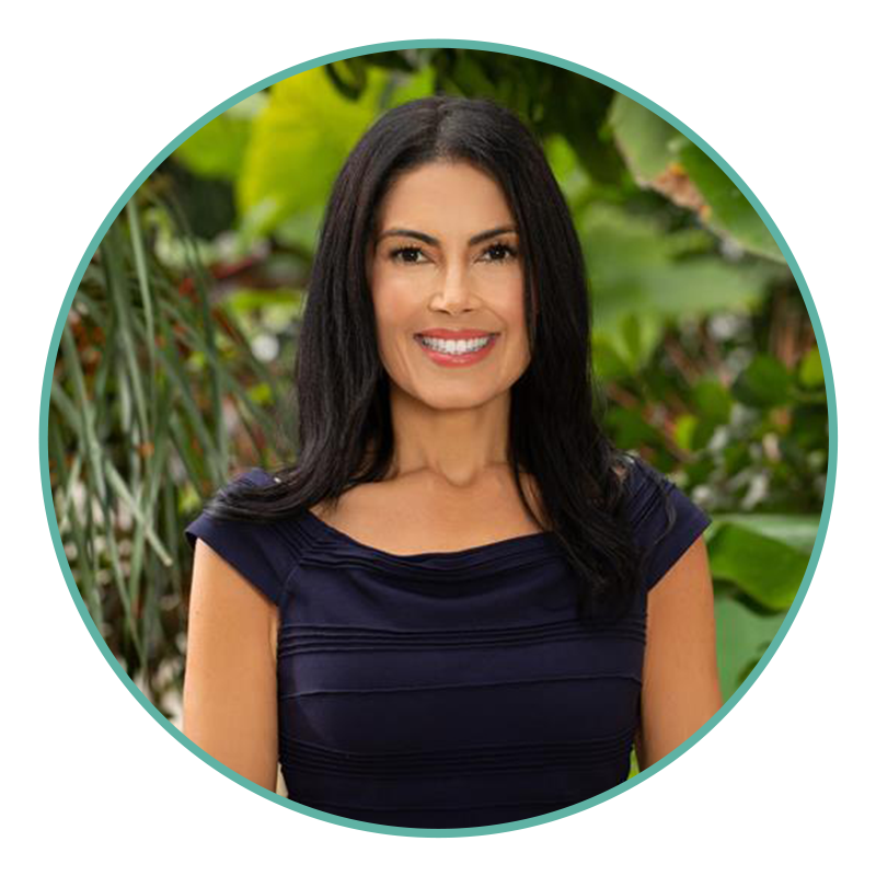 Woman with long dark hair wearing a navy blue top, standing outdoors in front of green leafy plants, smiling at the camera.