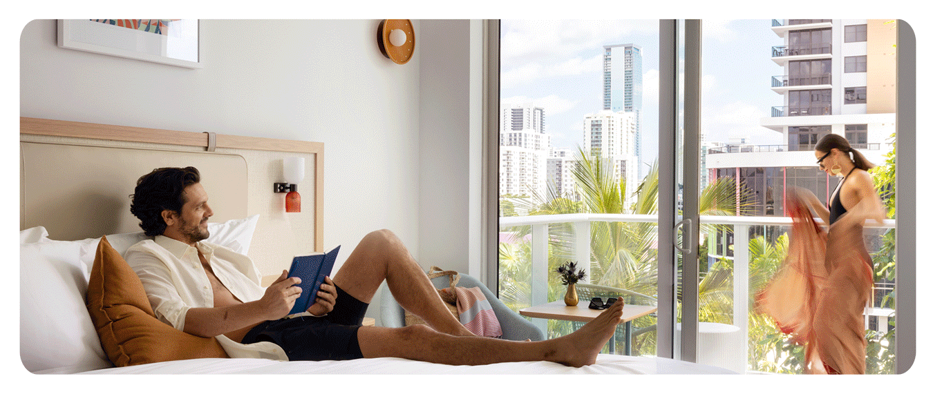 A man sits on a hotel bed reading a book while a woman steps onto a balcony; city buildings and palm trees are visible outside.